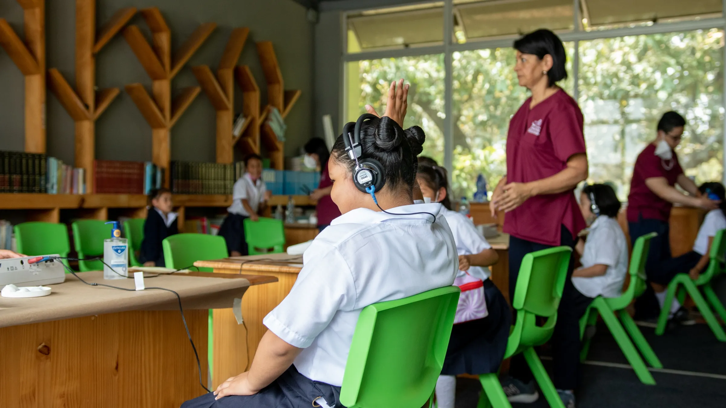 A young student in the foreground, facing away from the camera, raises their hand during a hearing screening test. They are wearing headphones hooked up to a testing device. In the background, aides assist other students with the screening test.