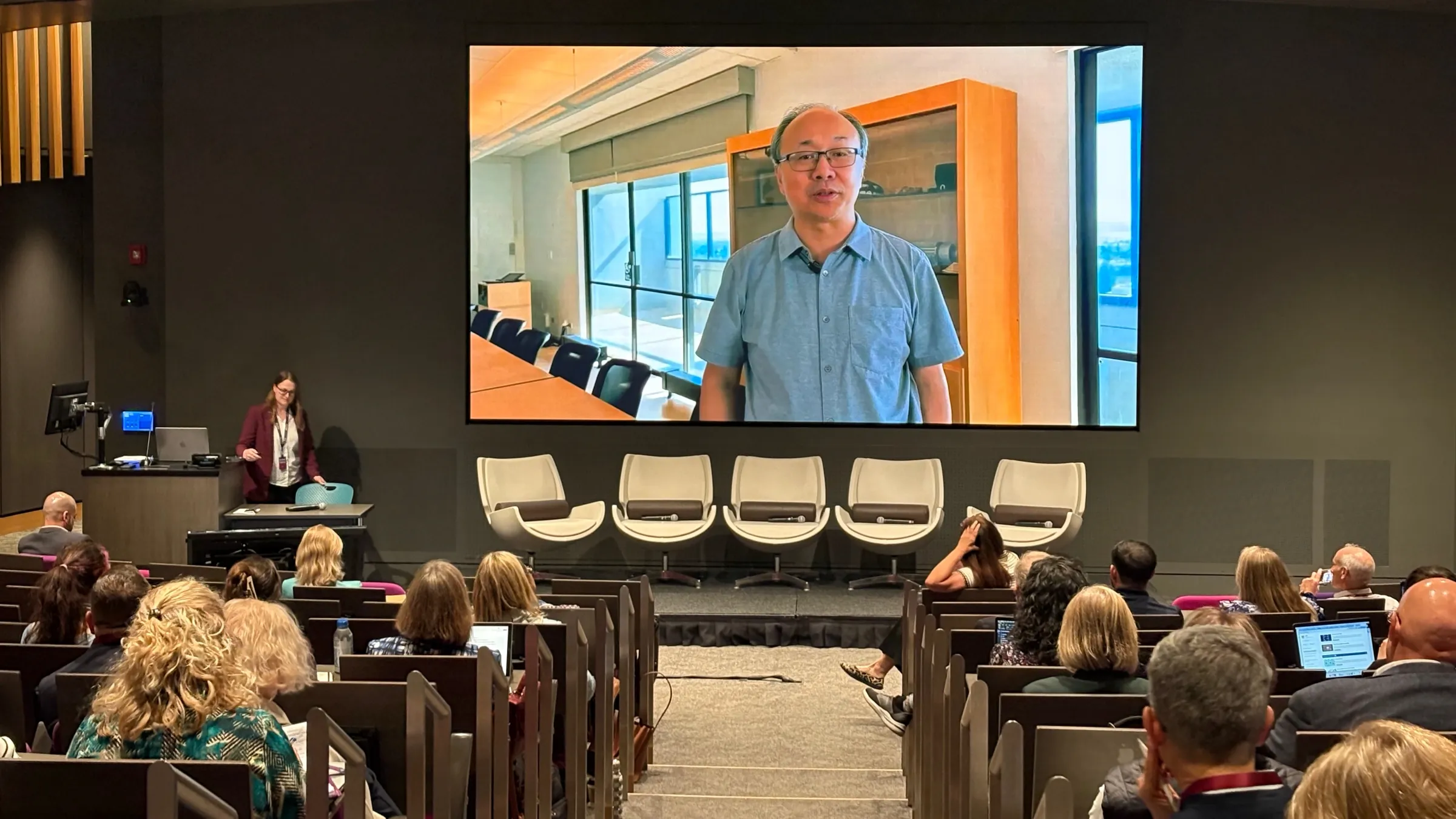 Ruth Steyn presents to a crowd in an auditorium at the AIxEd Leadership Summit, assisted by Professor Xiao-Li Meng's AI avatar displayed on a screen at the front of the room.