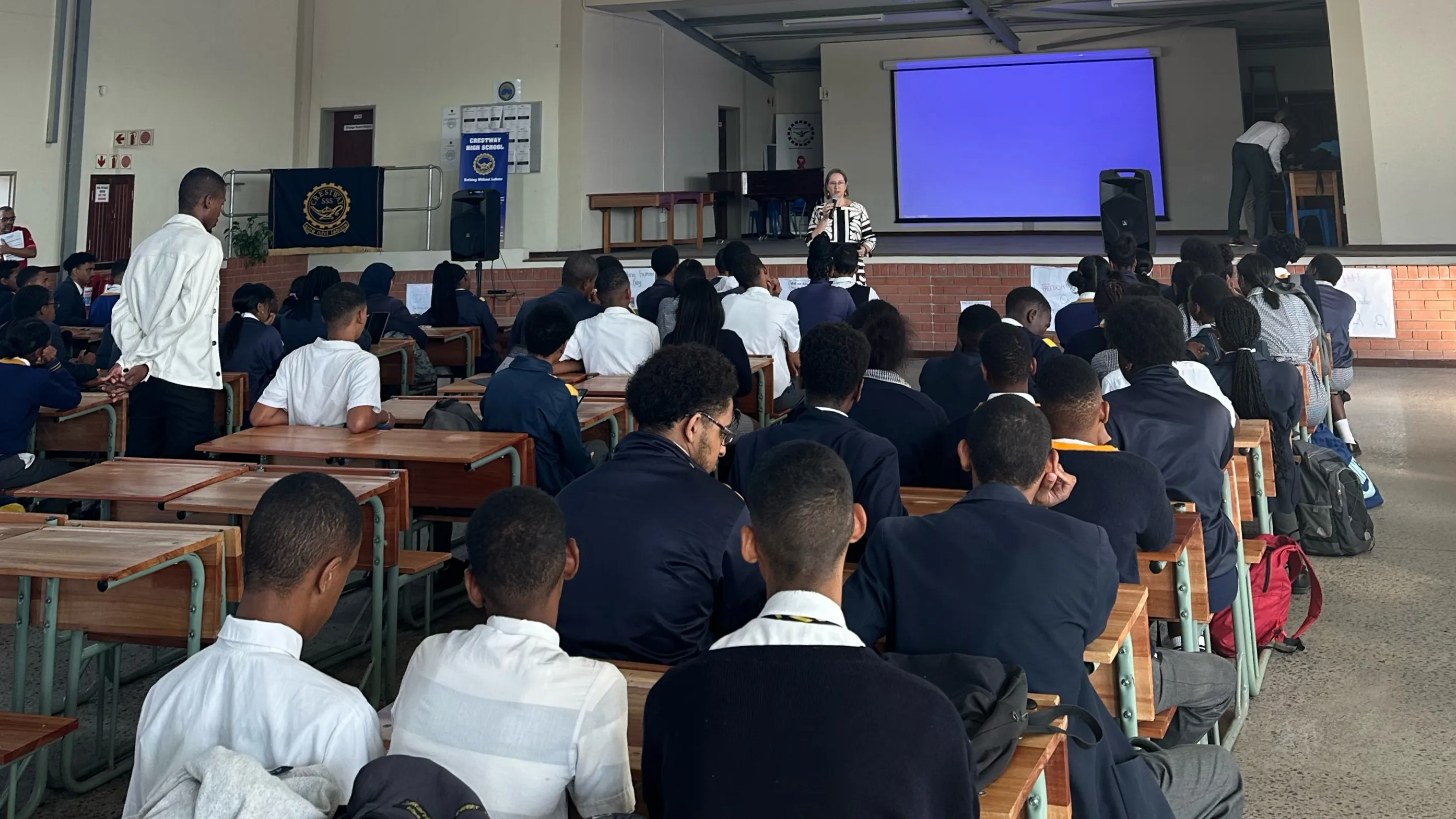 Students in school uniforms sit at wooden desks facing educator Nicole Masureik, who is speaking at the front of a large hall with a blue projection screen behind her, at Crestway High School.