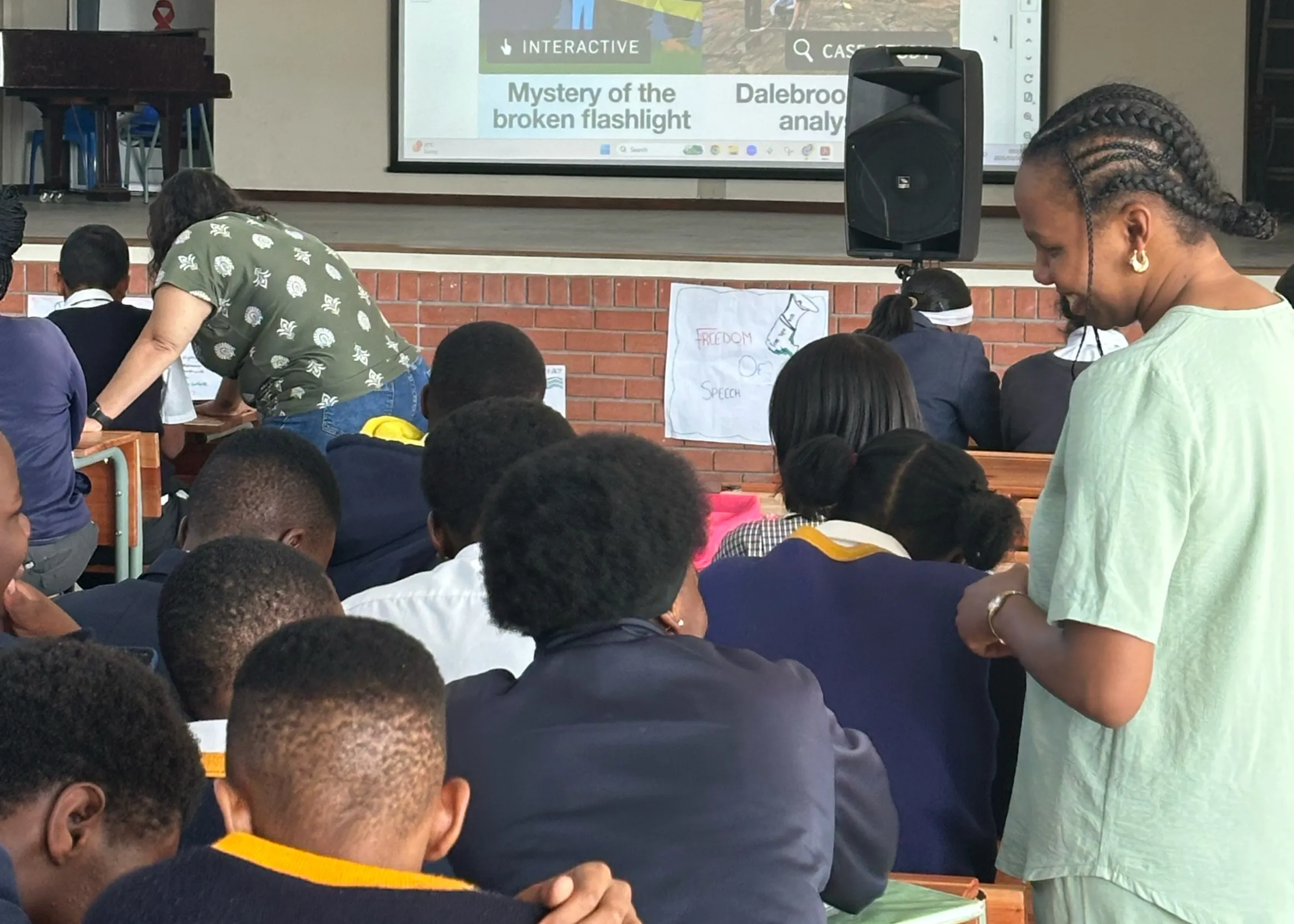 Students in school uniforms work at desks while two teachers assist them. A projected screen displays "Mystery of the broken flashlight" interactive lesson.
