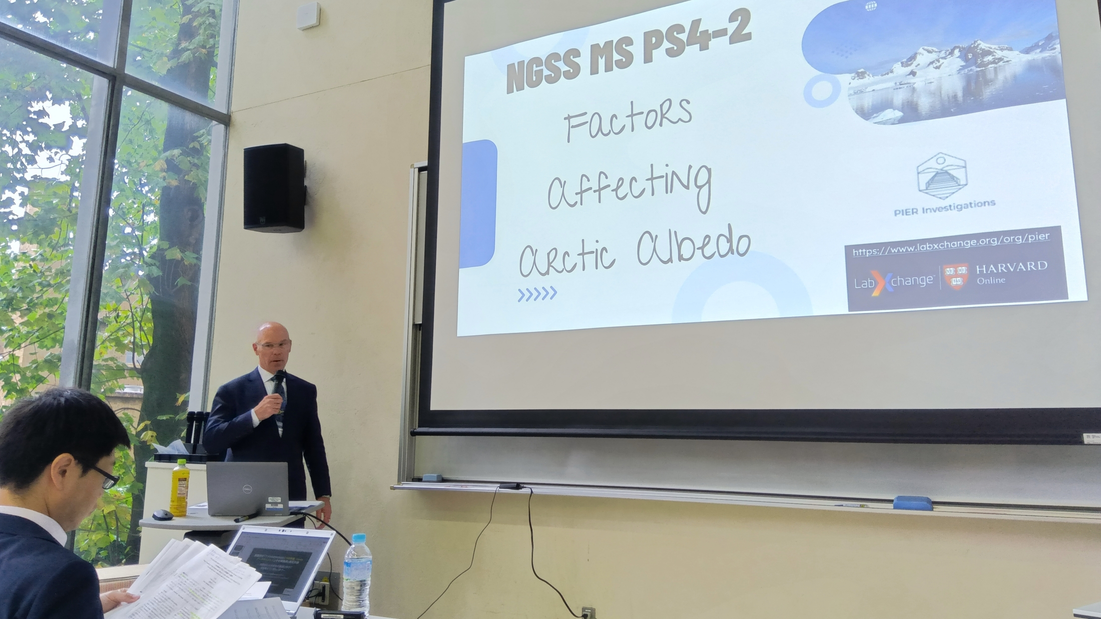 A presenter, Kenneth Huff, speaks at a podium beside a projected slide titled "NGSS MS PS4-2: Factors Affecting Arctic Albedo" with Harvard Online and LabXchange logos.