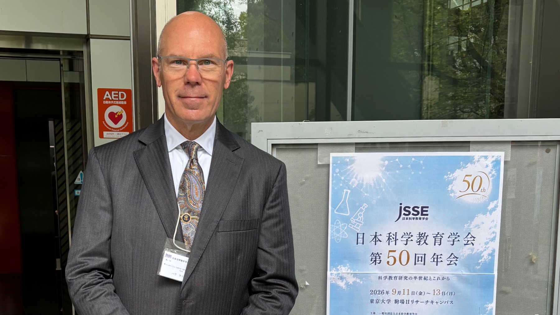 A man (Kenneth Huff) in a gray suit and paisley tie standing beside a JSSE 50th annual conference poster written in Japanese, outside a building.