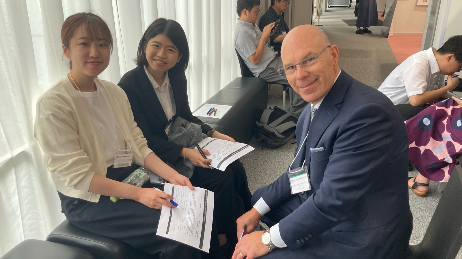 Kenneth Huff (right) and two conference attendees sit together in a hallway, smiling at the camera while reviewing printed documents.