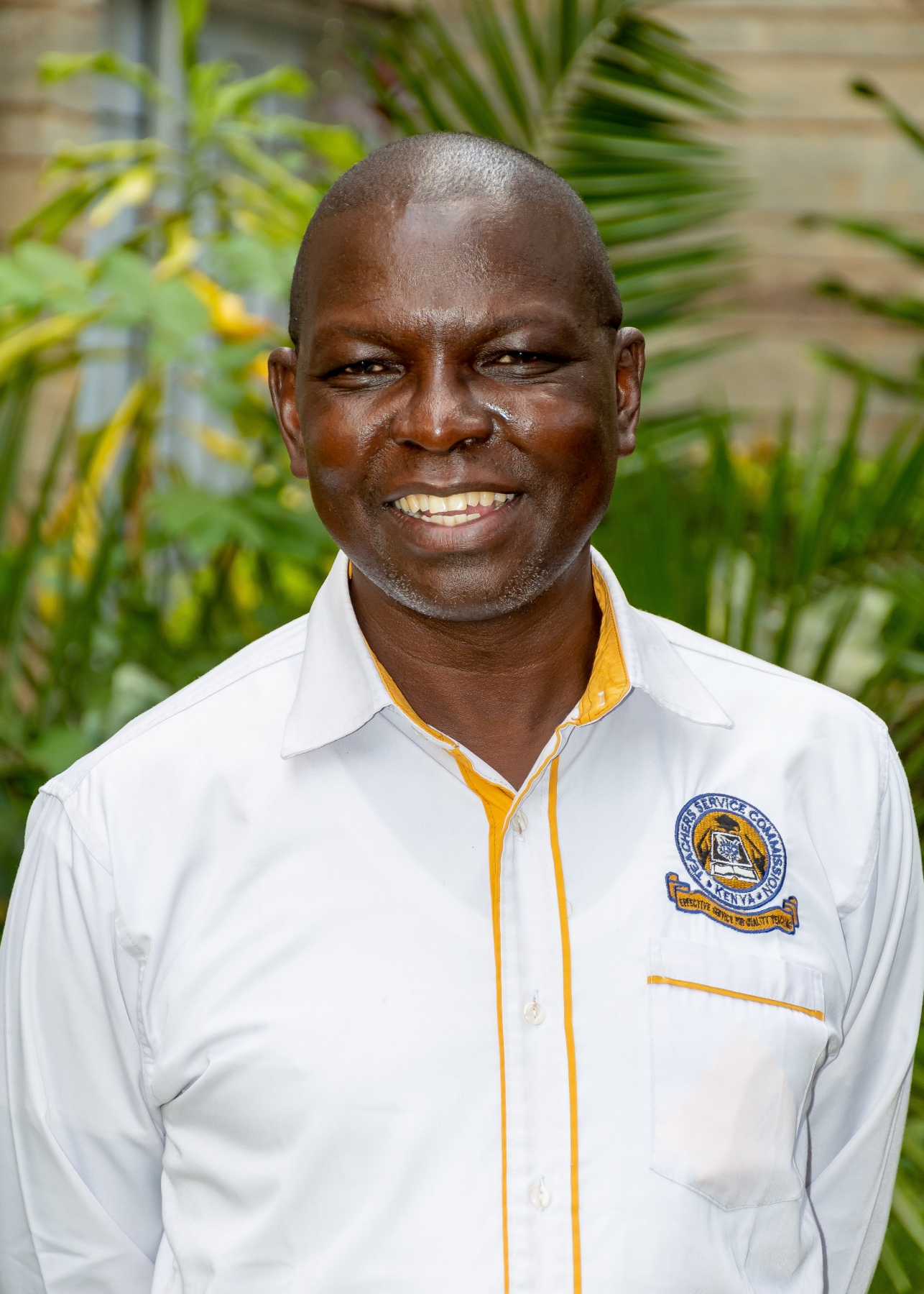 Smiling man wearing a white uniform shirt with the Teachers Service Commission Kenya emblem, standing outdoors near green tropical plants.