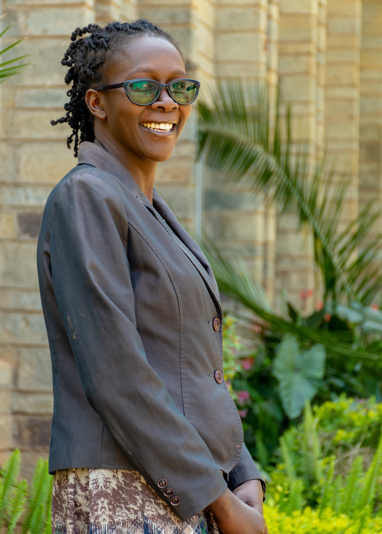 A smiling woman with twist locs and glasses wearing a gray blazer, standing outdoors near tropical plants and a stone building.