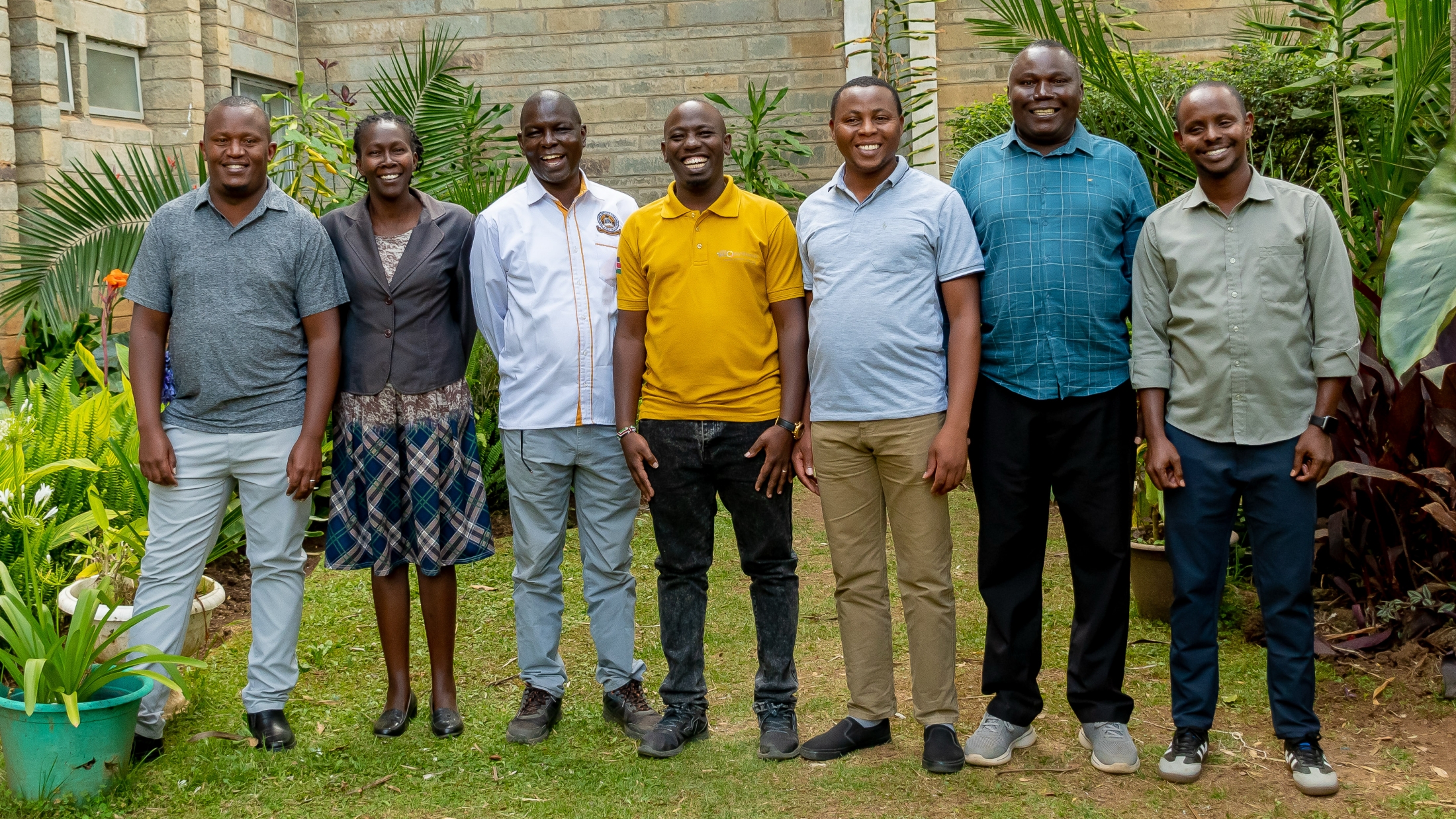 Seven smiling colleagues posing together outdoors on grass, with tropical plants and a brick building in the background.