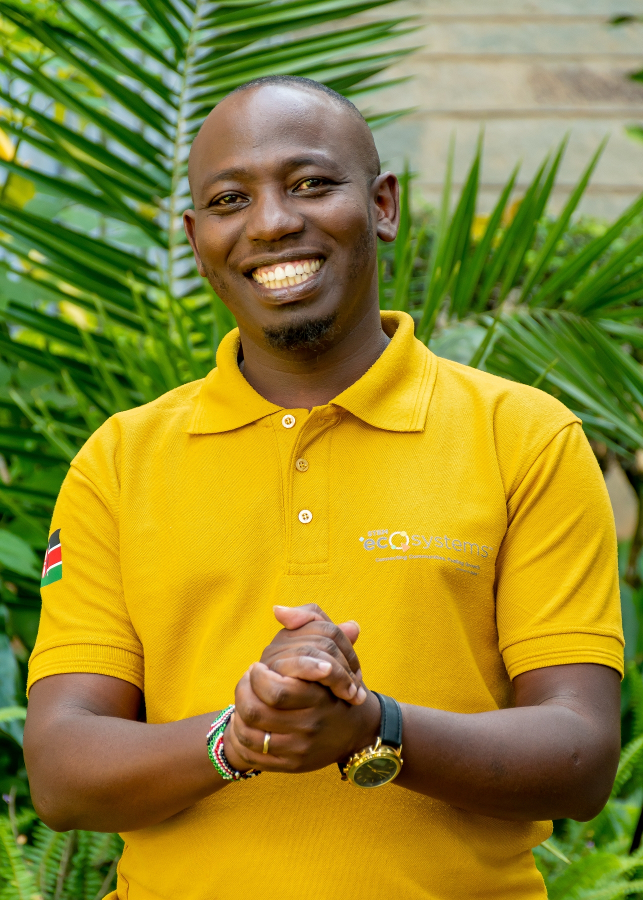 Smiling man wearing a yellow STEM ecOsystems polo shirt with a Kenyan flag patch, hands clasped, standing before lush green foliage.