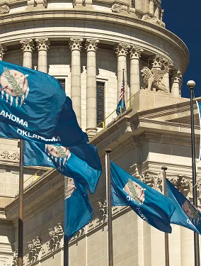 Oklahoma state flags waving in front of a classical building with columns and a lion statue under a clear blue sky.