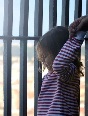 Young girl with curly hair in a striped sweater standing near a window with vertical bars, adjusting her sleeve.