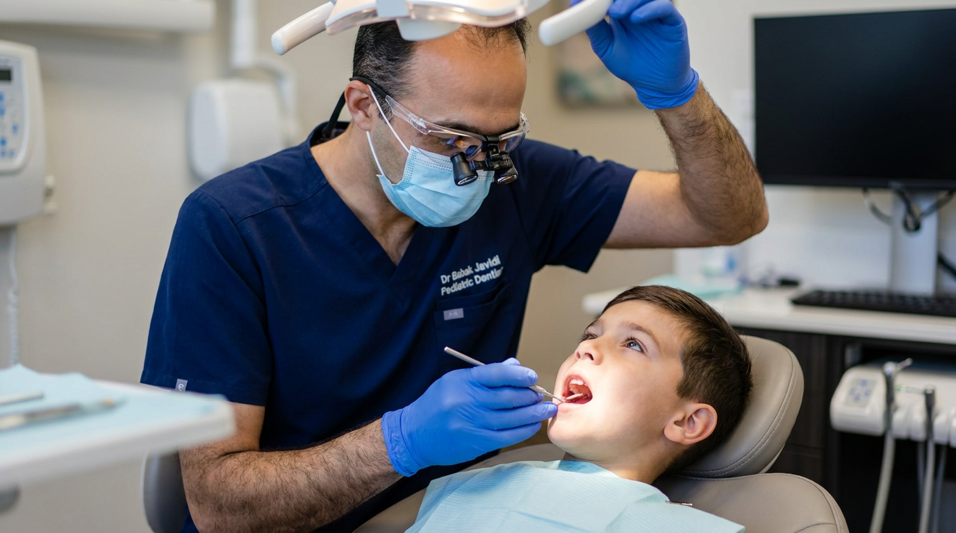 Dr. Javidi, wearing blue scrubs, a mask, and loupes, examines a boy in a dental chair with a dental mirror and light.