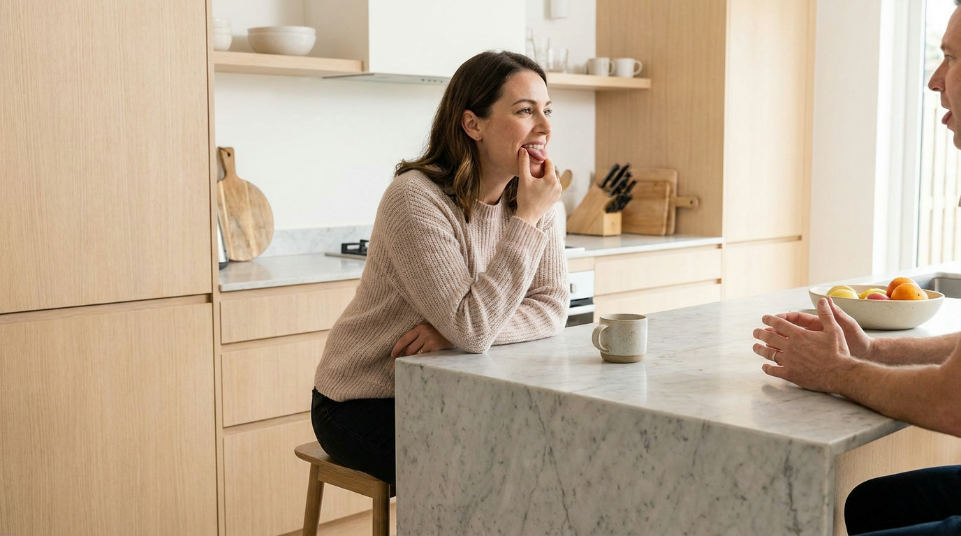 Samantha sits on a stool at a marble kitchen island, wearing a light pink sweater and black pants, looking right with her finger touching her mouth. A mug is on the island, and a man's hands are visible on the far side of the island. The