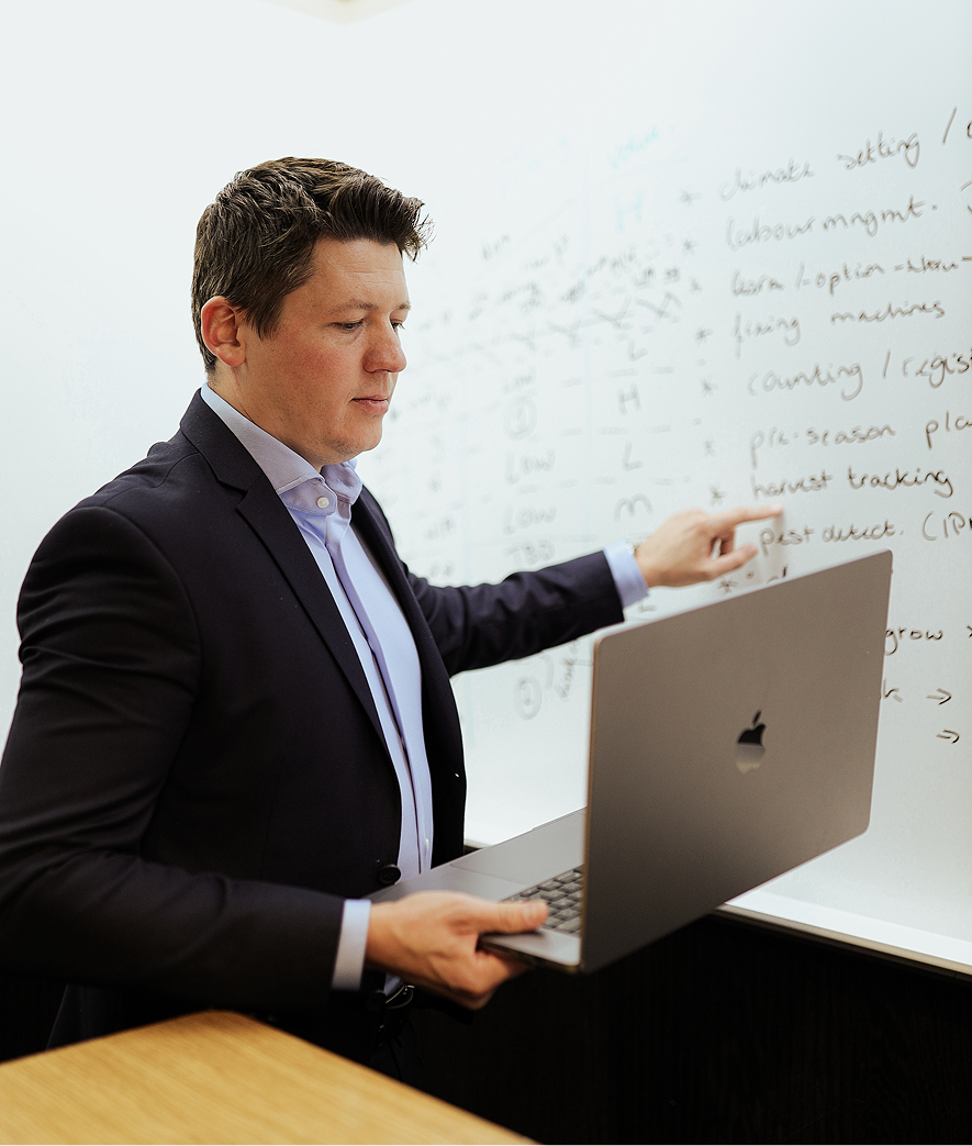 Alex Kukharenko, PixelDarts founder, in a dark blazer holding a laptop and pointing at handwritten notes on a whiteboard.