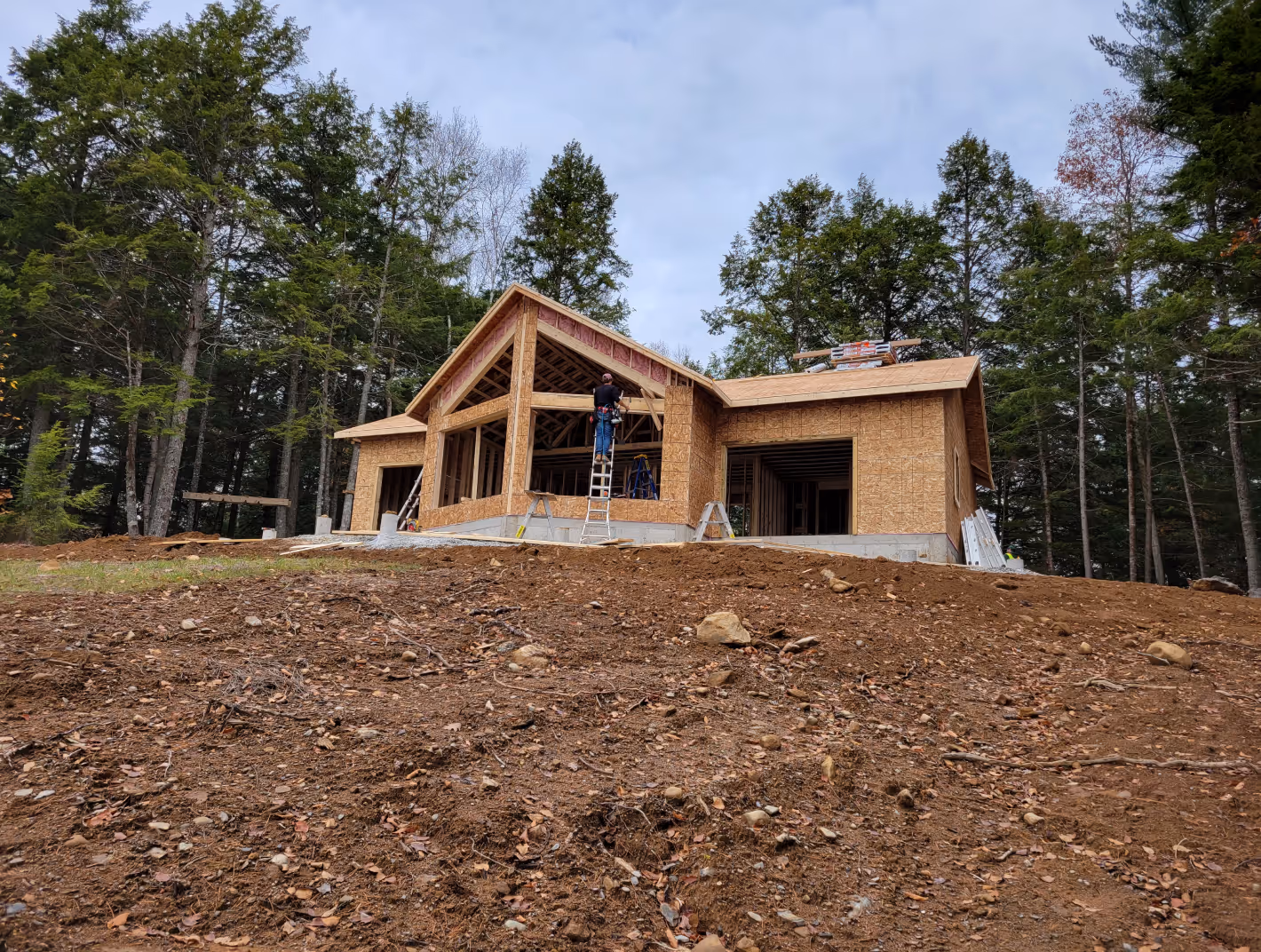 Home builders in halifax nova scotia - New house under construction on a cleared, rocky dirt lot surrounded by tall evergreen trees. Home builder in Halifax, Nova Scotia.