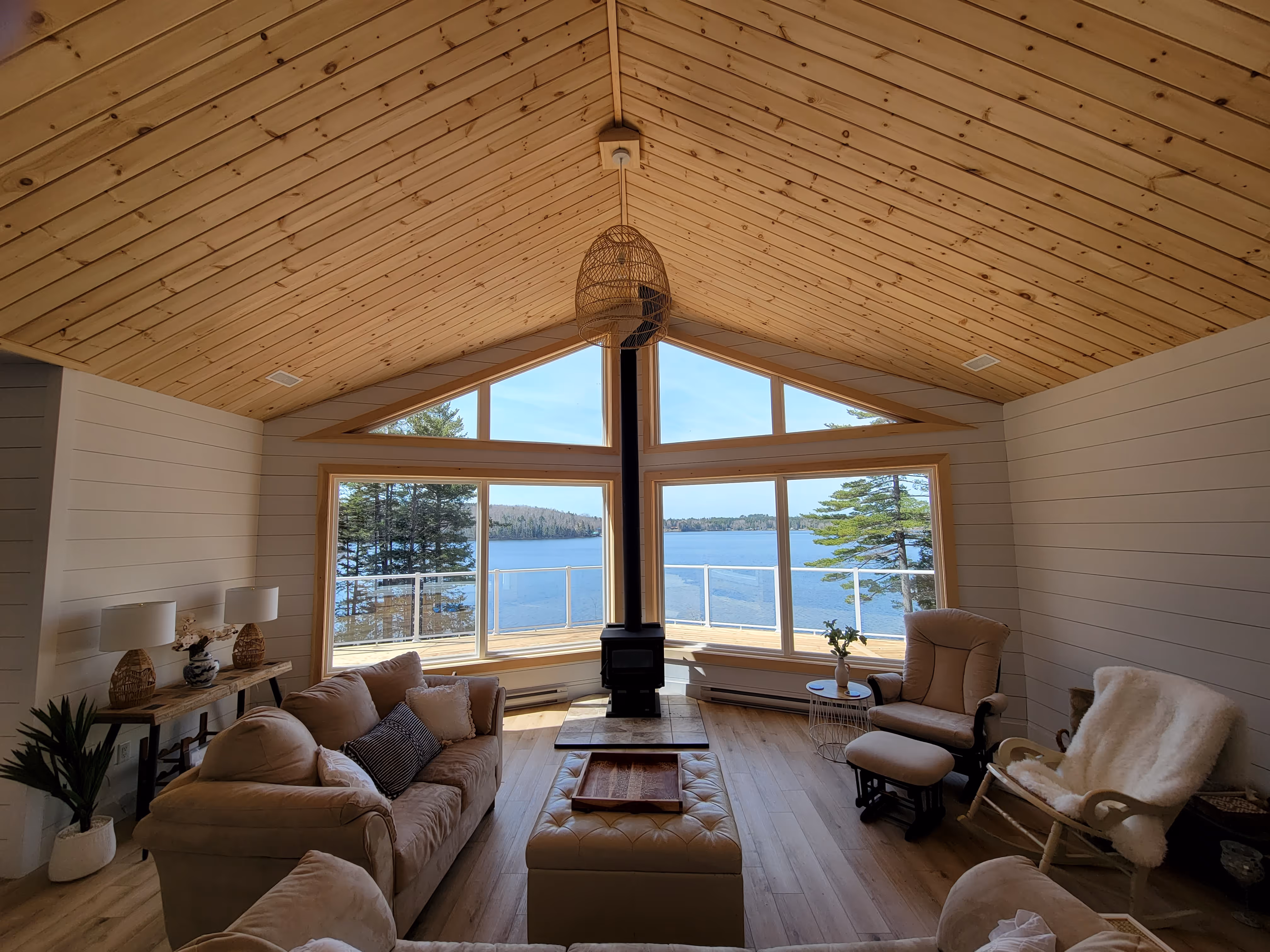 Residential home builders- Cozy living room with beige sofas, a rocking chair with a white throw, a wood stove, and large windows overlooking a lake and trees. New construction in Halifax, Nova Scotia.