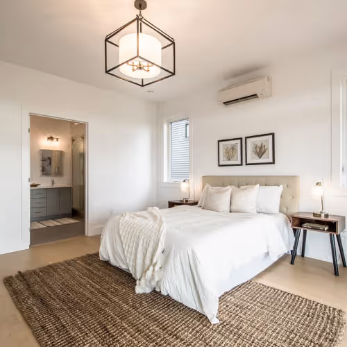 House builders in nova scotia -Bright modern bedroom with a white bed, two bedside tables with lamps, a large woven rug, wall art, and an open doorway to an ensuite bathroom. Home builder in Halifax, Nova Scotia.