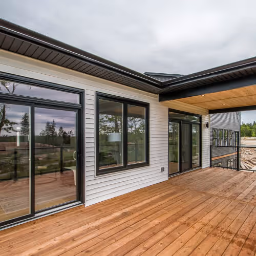 Home builders ns -Modern house exterior with large glass sliding doors and windows opening onto a spacious wooden deck under a cloudy sky. Home builder in Halifax, Nova Scotia.
