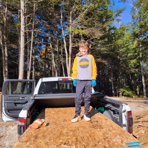 home contractors near me - Young boy wearing a yellow and gray hoodie, blue gloves, and dark pants standing on a wooden board in the bed of a white pickup truck with an open door, surrounded by a forest.