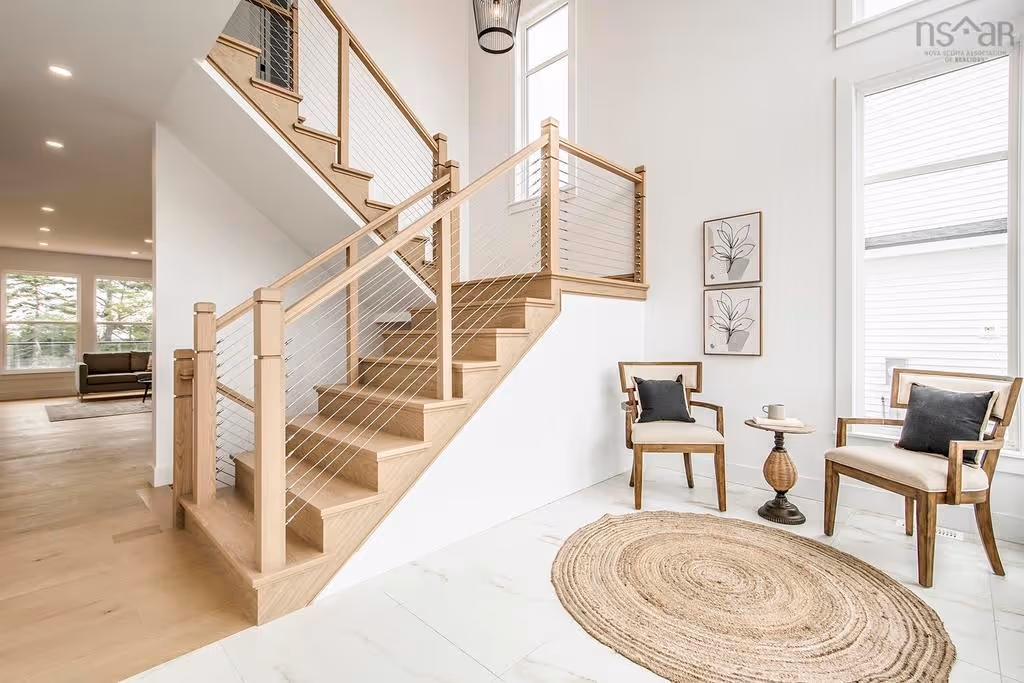 Signature homes halifax -Modern entryway featuring light wood staircase with cable railing, two wooden chairs with dark cushions, round woven rug, and small wooden side table.