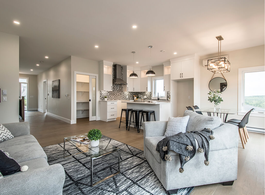 Modern open-concept living room and kitchen with gray sofas, glass coffee table, white cabinetry, and a dining area with black chairs and a large window. Signature Home Builders in Halifax, Nova Scotia