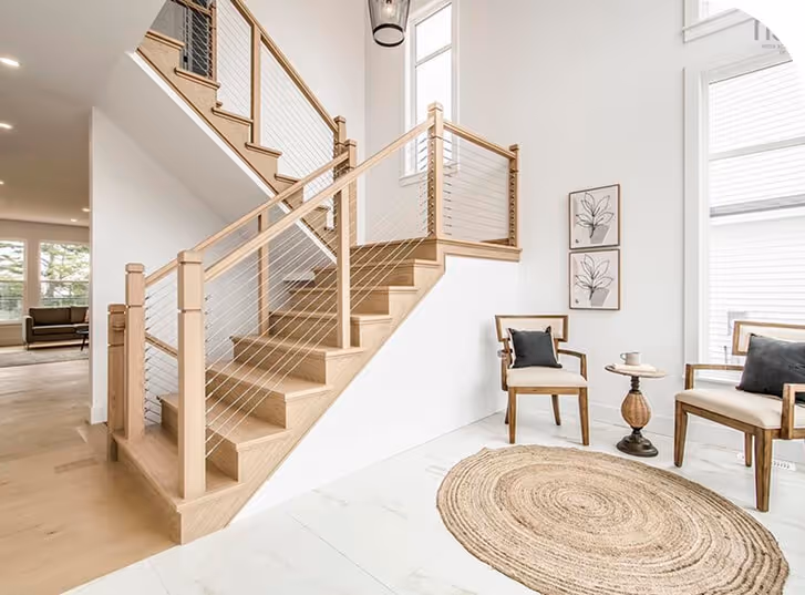 Bright modern entryway with wooden staircase and cable railing, white walls, round jute rug, two accent chairs, and large windows letting in natural light.