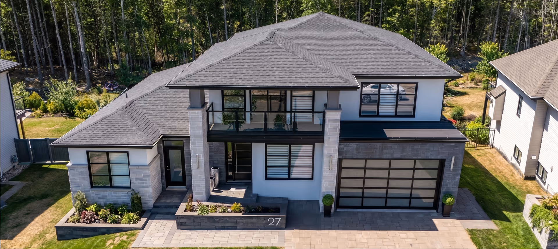Aerial view of a custom-built home in Nova Scotia featuring a dark shingle roof, large black-trimmed windows, stone and stucco exterior, and attached garage surrounded by mature trees