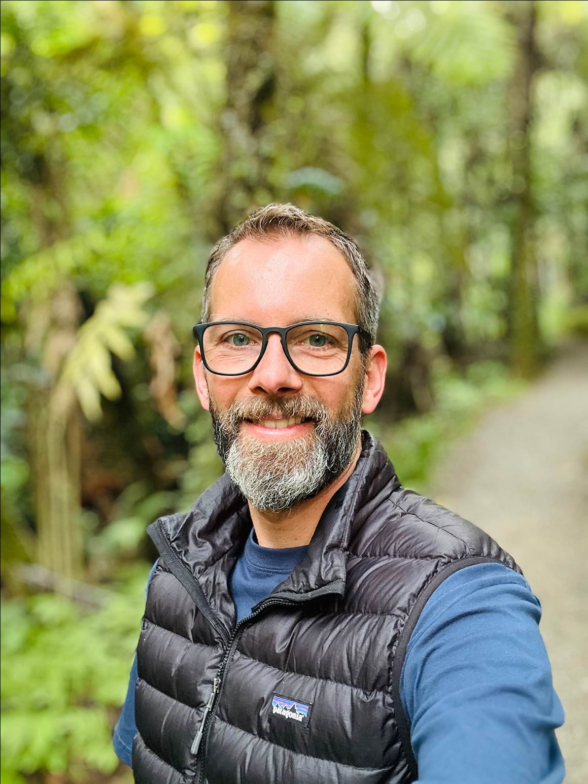 Portrait of Gunnar Froh standing outdoors in soft natural light. He has short dark hair, a neatly trimmed beard, and is wearing round glasses. He is smiling warmly and dressed in a dark jacket over a white shirt. The background is blurred, with greenery and a hint of sky, creating a calm and approachable atmosphere.