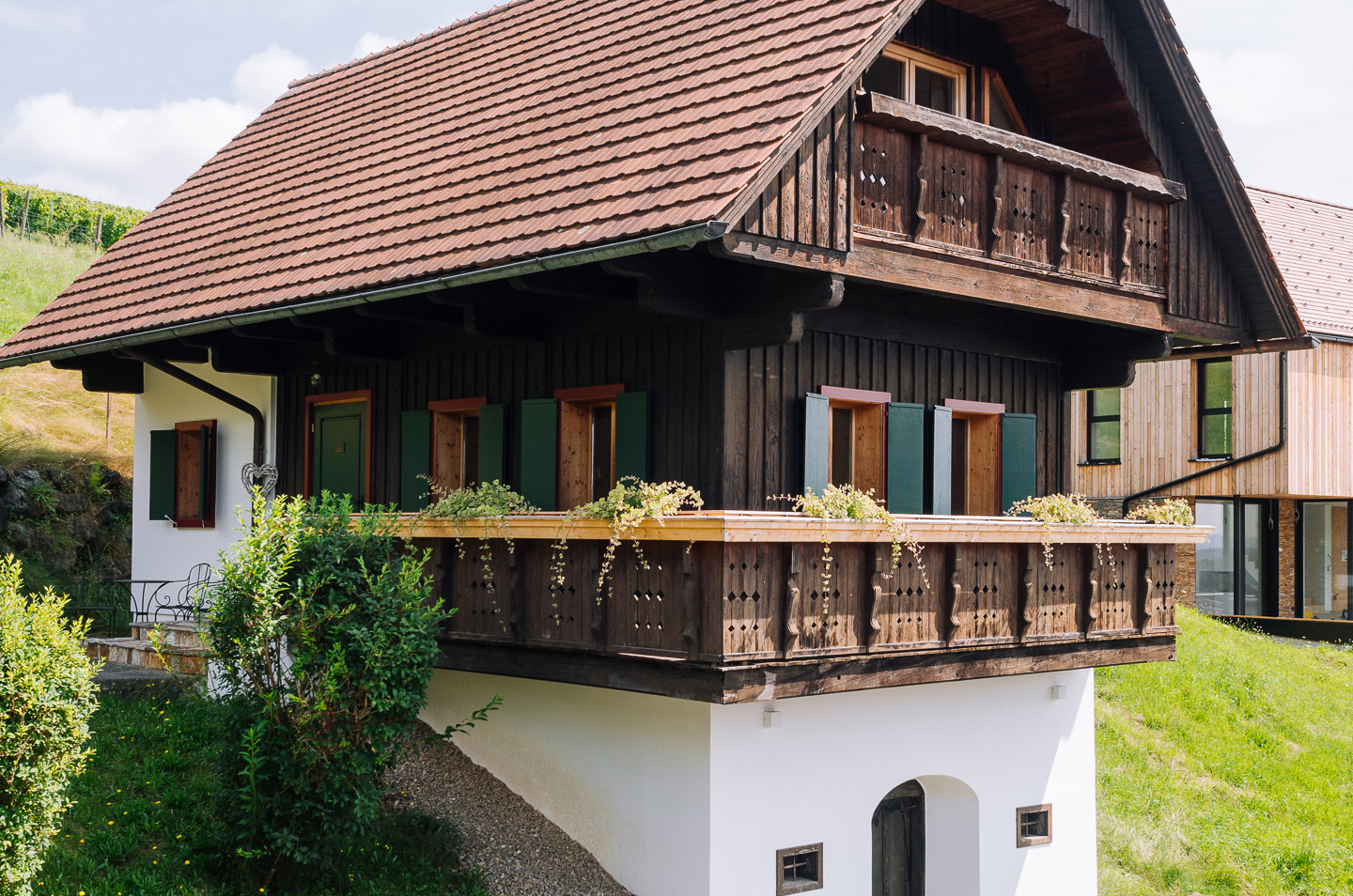 Holzhaus mit rotem Ziegeldach, umgeben von grünem Garten mit Rosen und einem Treppenweg im Sonnenschein.