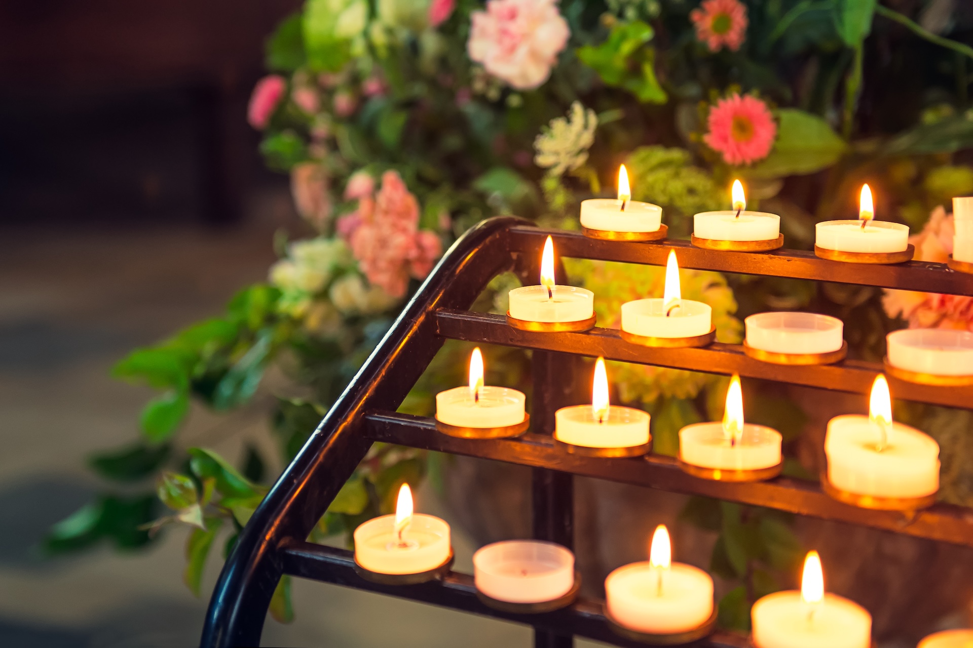 Lit tea light candles on a black metal rack with blurred pink and green flowers in the background.