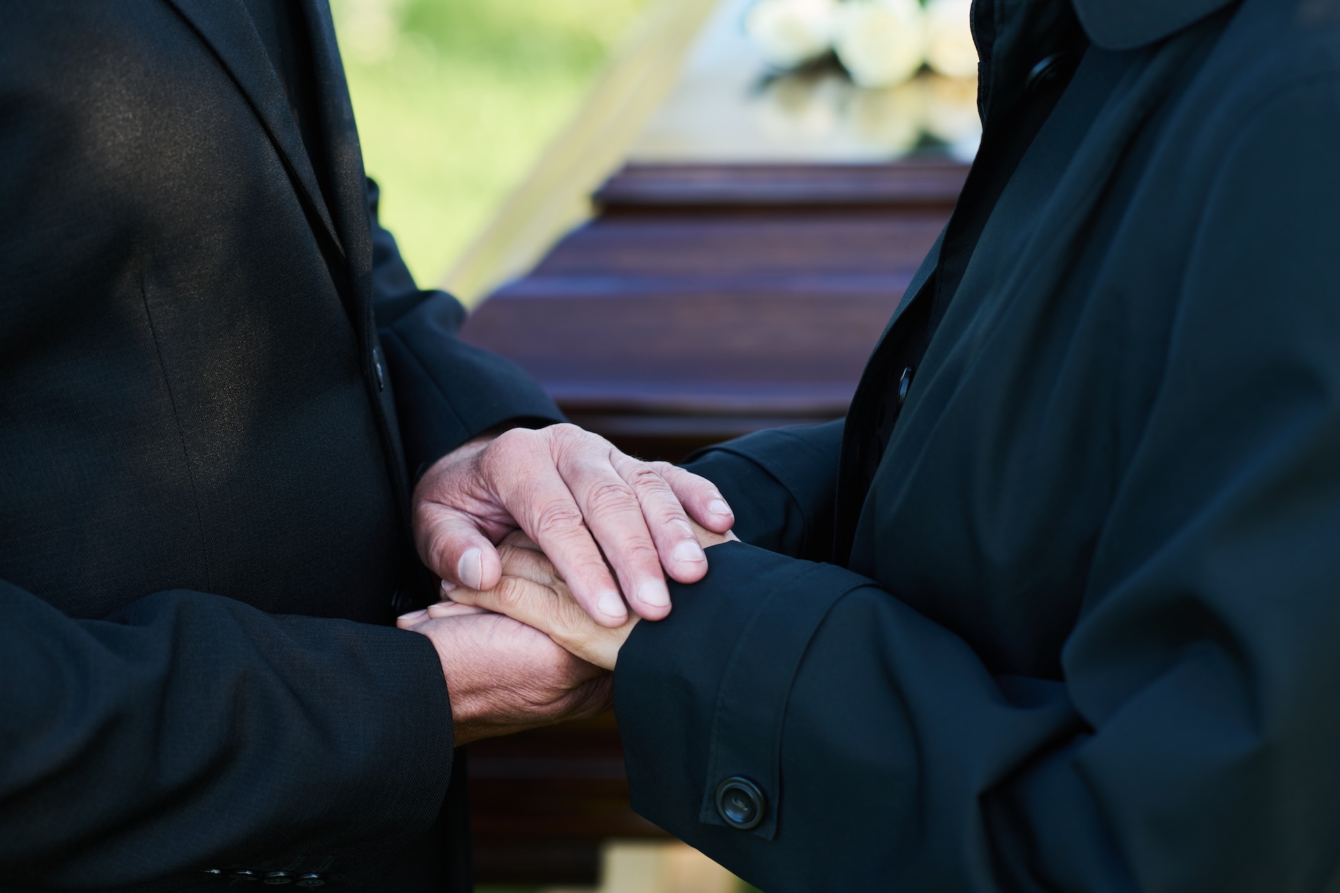Two people in black coats holding hands tightly in front of a wooden coffin at a funeral.