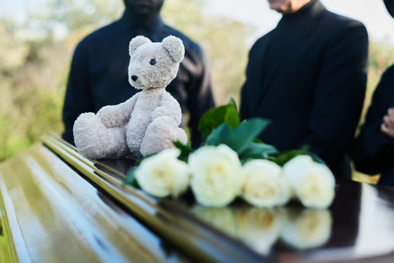 Teddy bear and white roses on a polished coffin at a funeral with mourners dressed in black.
