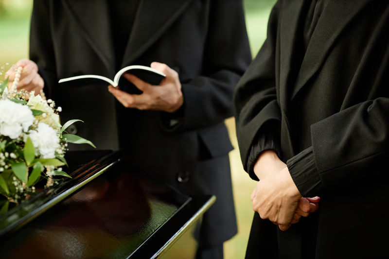 Two people in black coats at a graveside service, one holding a prayer book, the other with hands clasped.