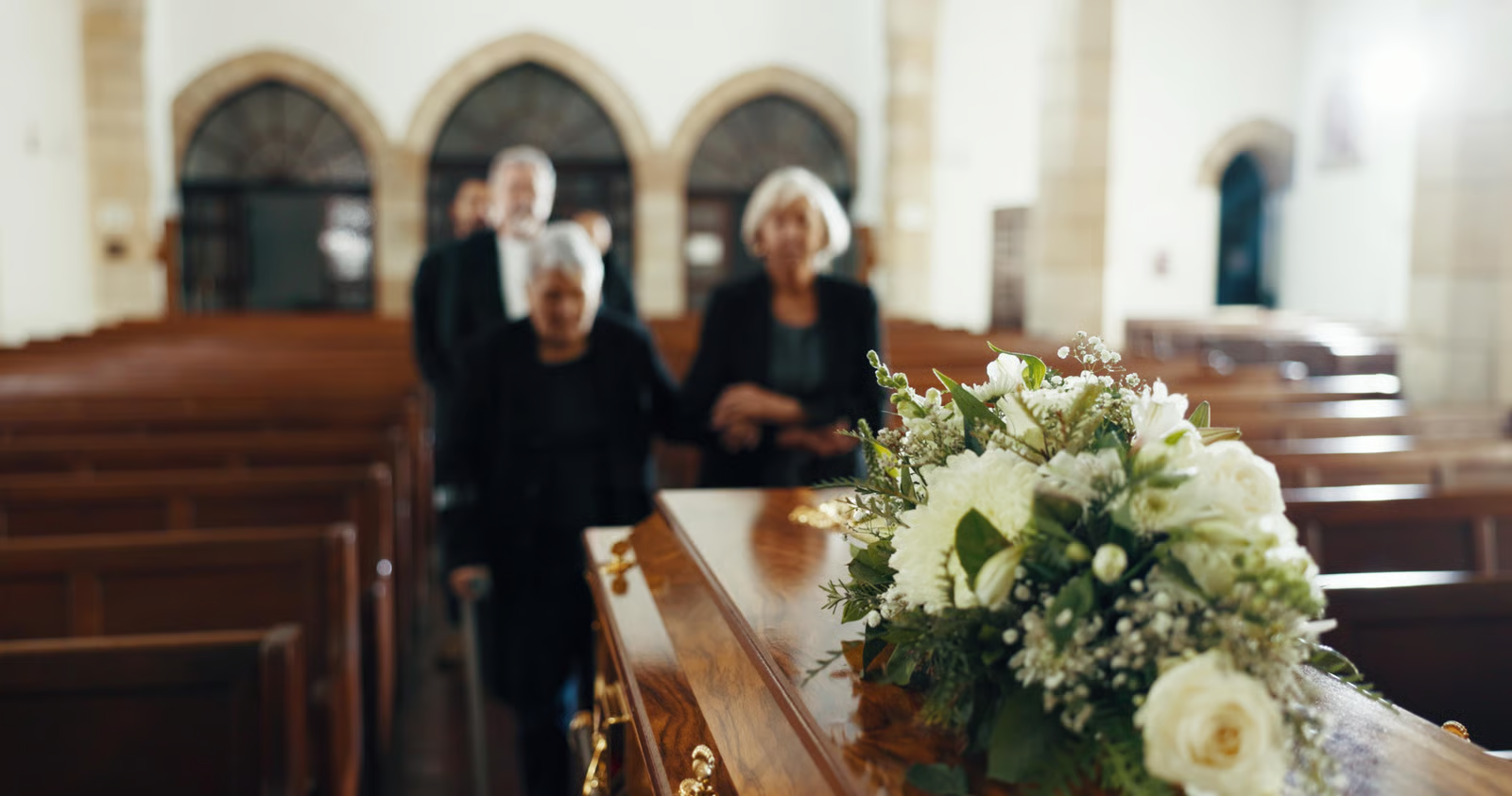 Close-up of floral arrangement on wooden coffin in church with mourners walking down aisle.