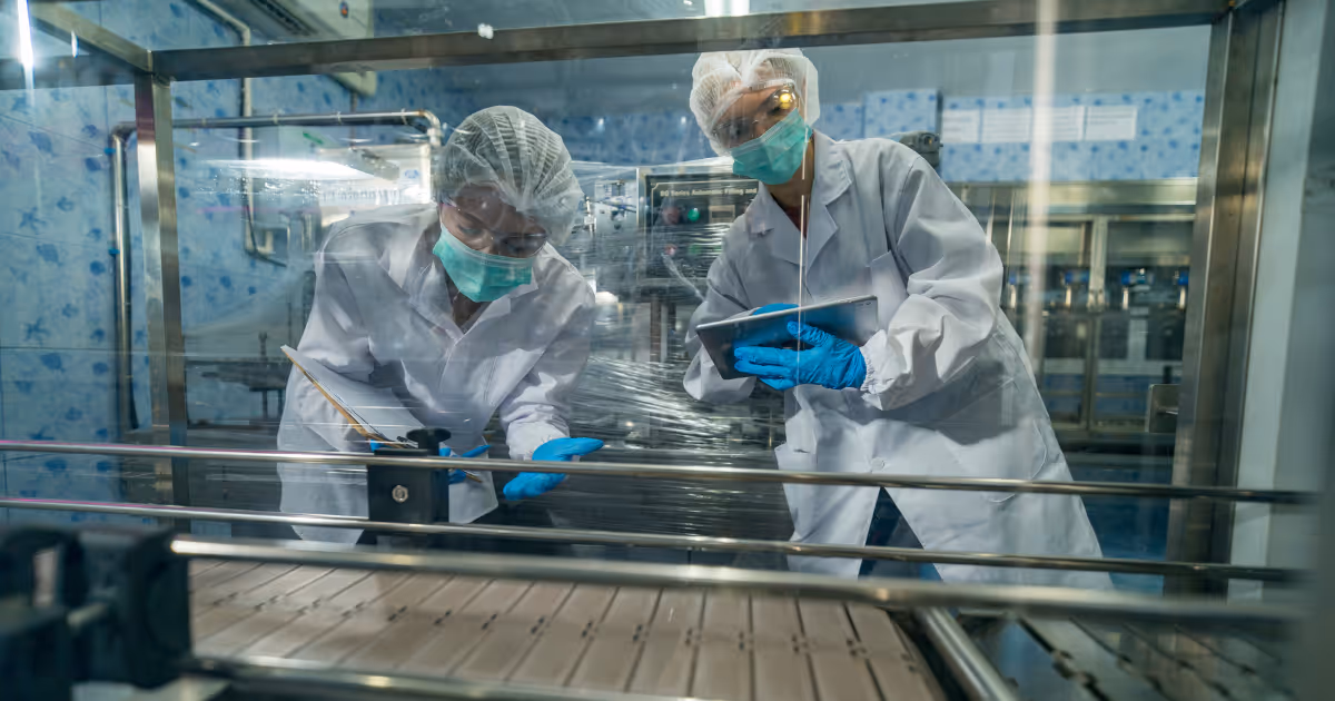 Two lab technicians wearing hairnets, masks, gloves, and white coats inspecting machinery in a clean industrial setting.