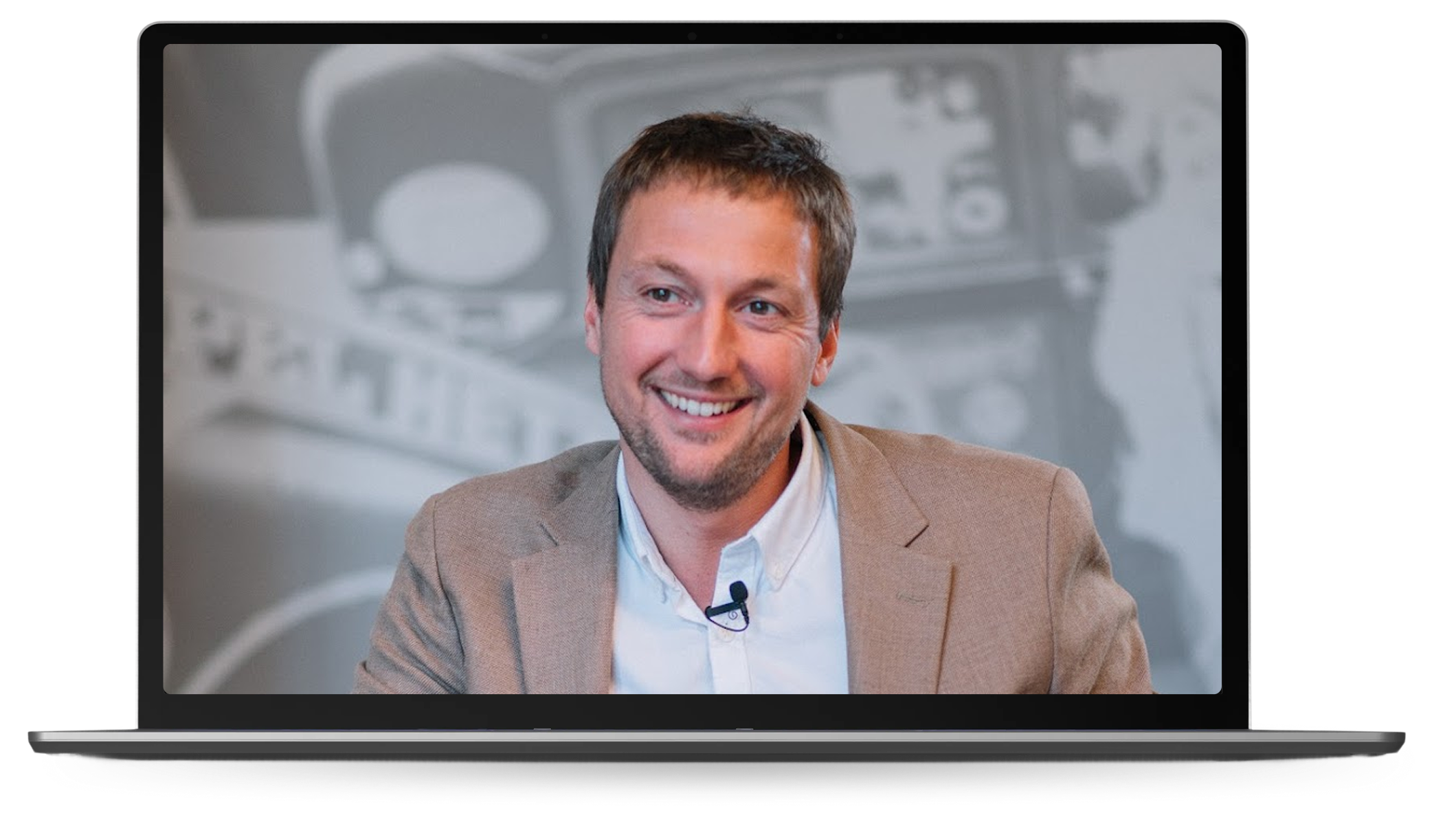 Smiling man wearing a brown blazer and white shirt speaking, shown on a laptop screen.