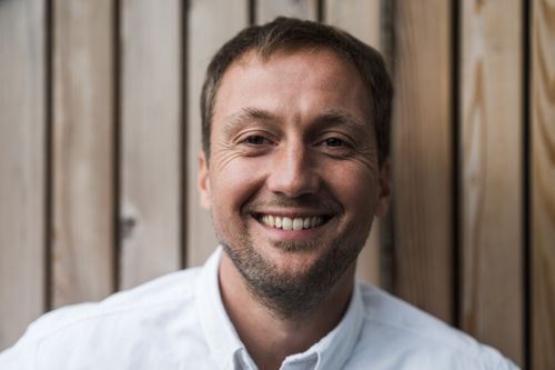 Smiling man with short brown hair and light stubble wearing a white shirt against a wooden panel background.