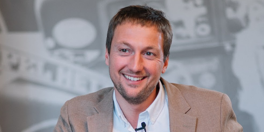Smiling man wearing a brown blazer and white shirt speaking, shown on a laptop screen.