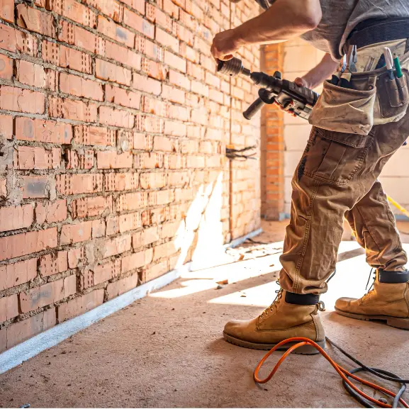 Worker using a power drill on an exposed brick wall in a construction site.
