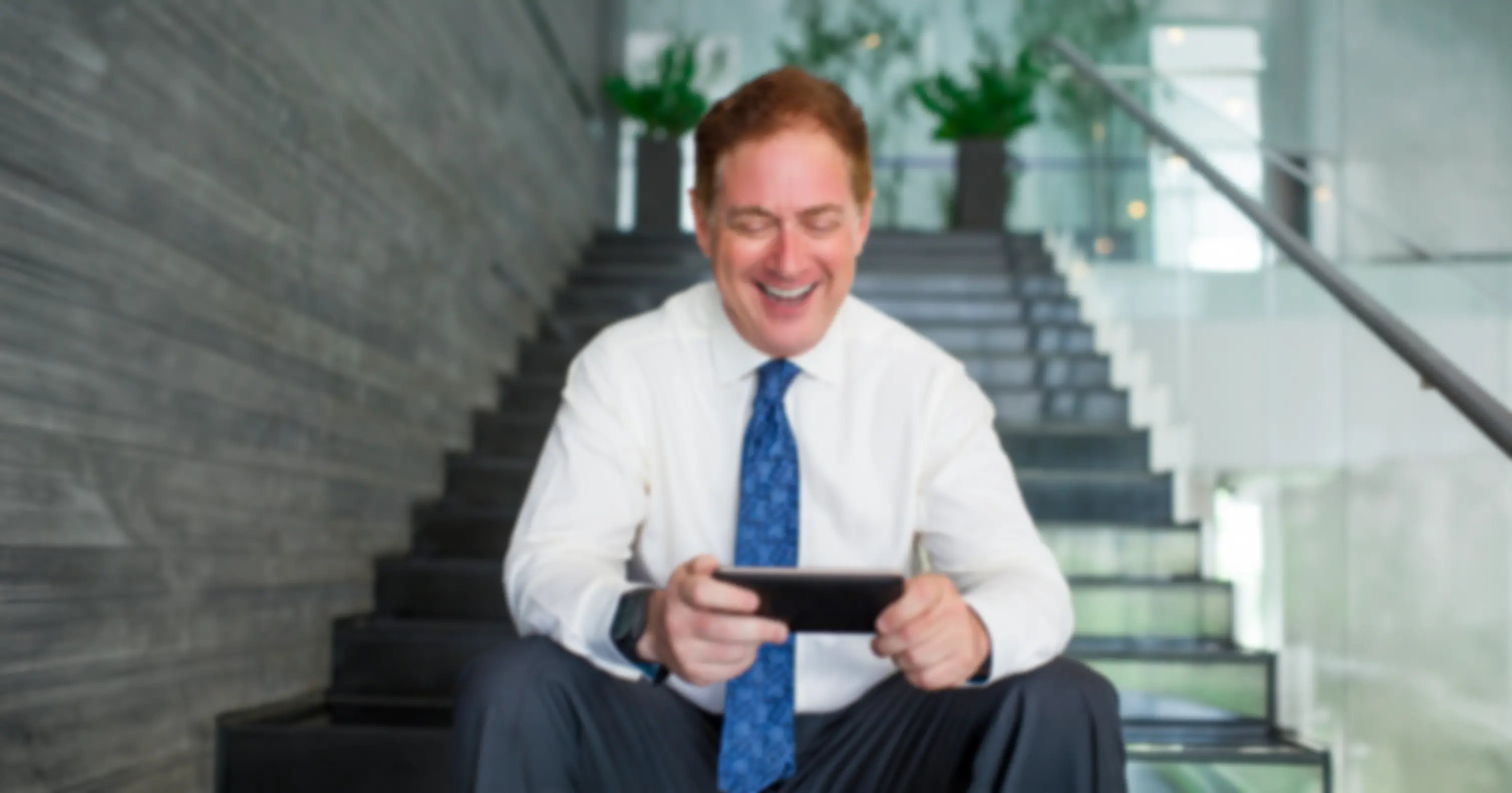Man in white shirt and blue tie sitting on stairs smiling while looking at smartphone.