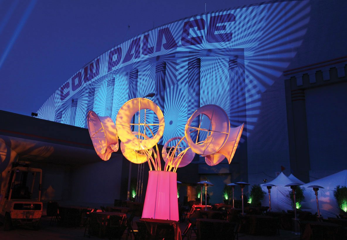 A giant blue gobo projection reads across the Cow Palace facade behind illuminated sculptural forms and an outdoor patio staged by Impact.