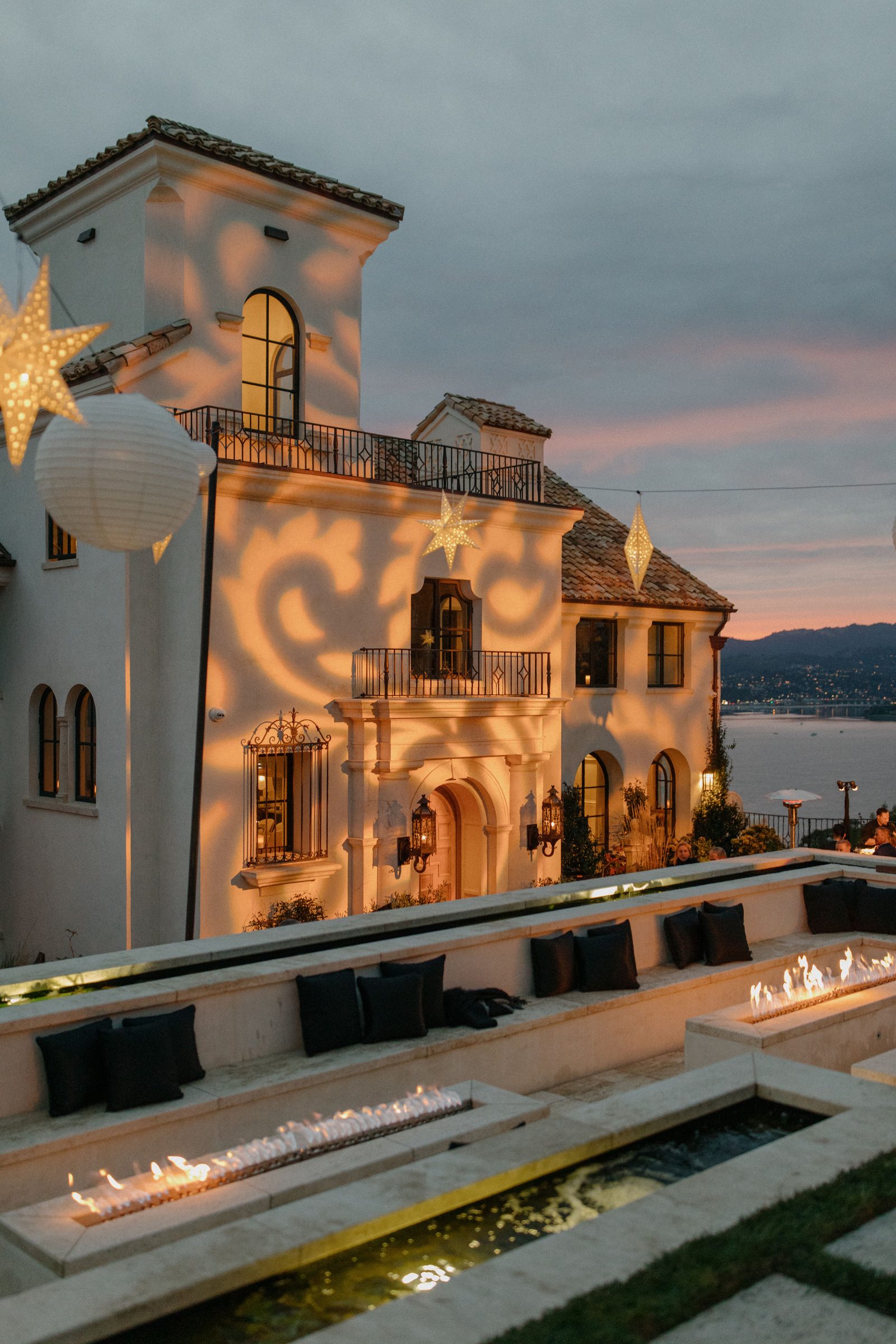 A Mediterranean hillside villa at dusk washed by Impact with decorative star gobos, glowing paper lanterns, and fire features overlooking the bay.