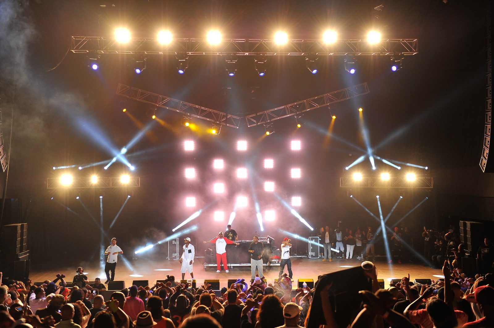 A hip-hop act performs on a large festival stage lit by Impact with intersecting aerial beams, an LED cube grid, and haze above a dense crowd.