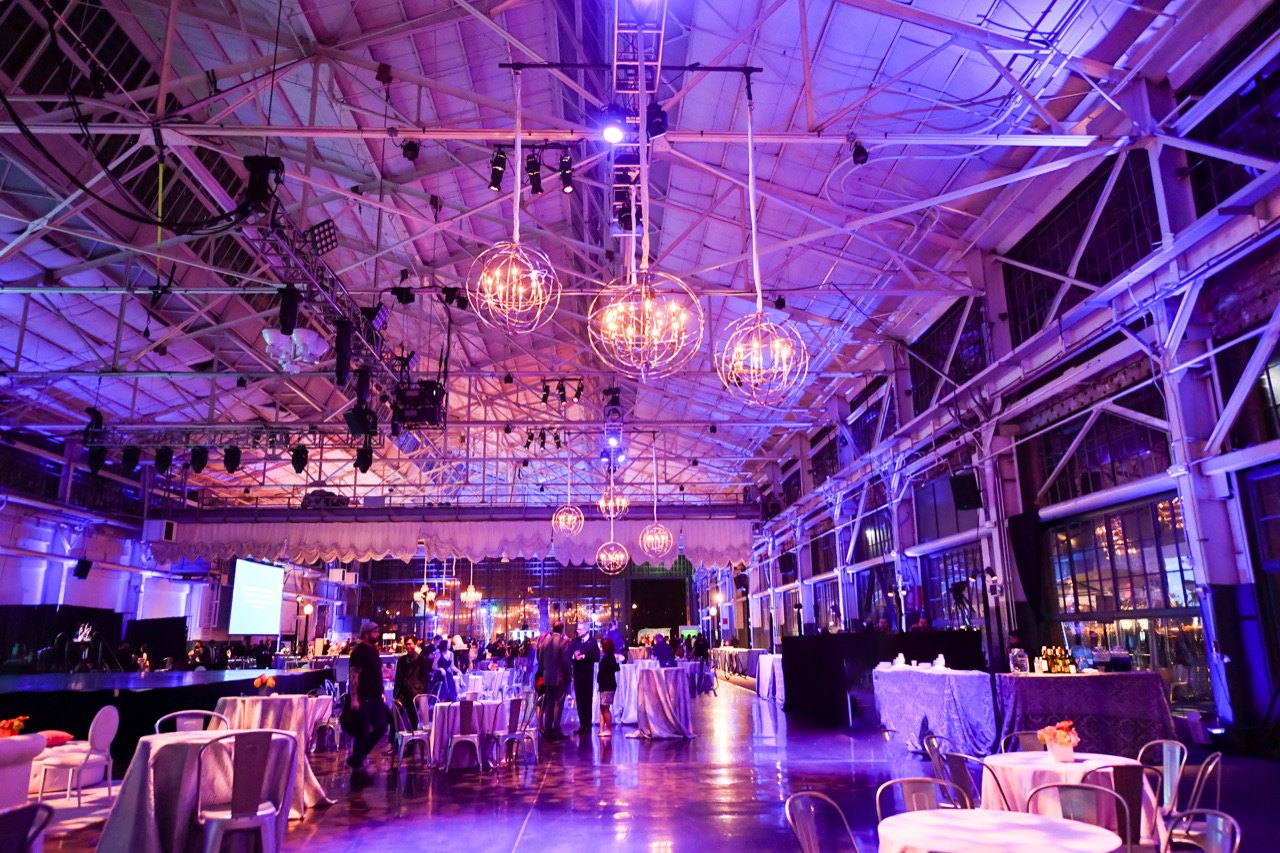 Impact bathes the industrial Craneway Pavilion in violet uplighting beneath suspended spherical orb chandeliers for a seated event.