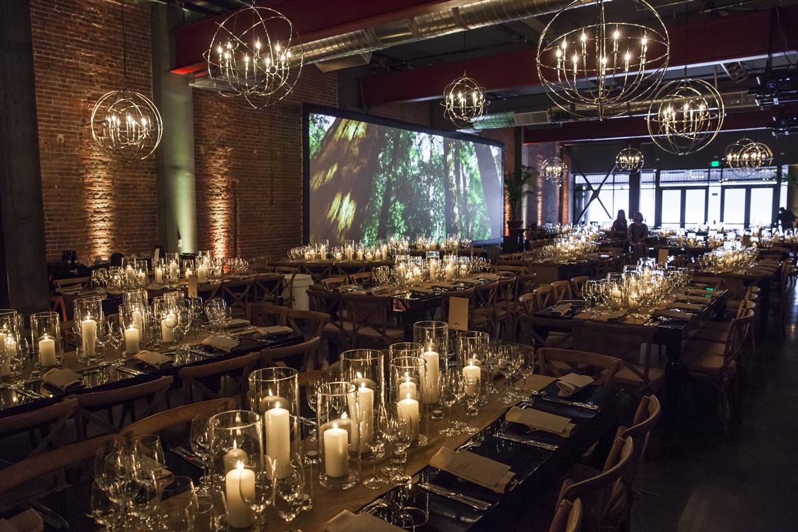 Candlelit long banquet tables run beneath Impact's silver orb chandeliers and a large forest projection on an exposed brick wall.