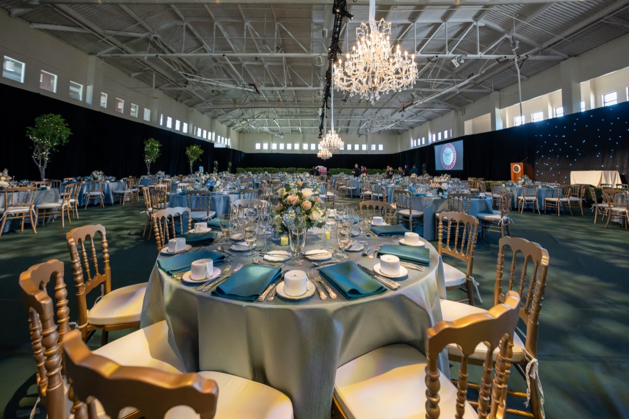 A crystal chandelier and twinkling star-curtain backdrop light Impact's formal gala dinner set with blue and gold table settings and gold chiavari chairs.