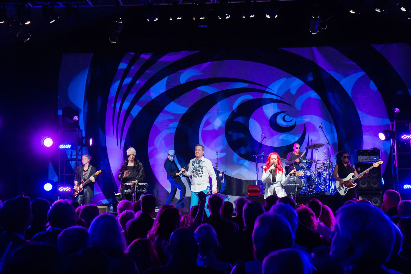 A live band performs before a packed audience under Impact's blue and magenta stage lighting and a swirling projected spiral backdrop.