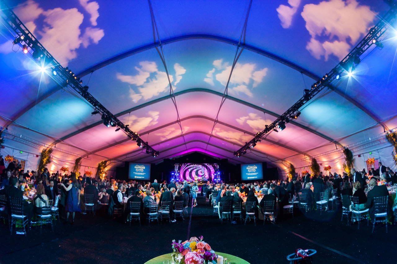 Impact projects a cloud-filled sky across a tent ceiling with stage lighting and warm uplighting at a hospital gala.