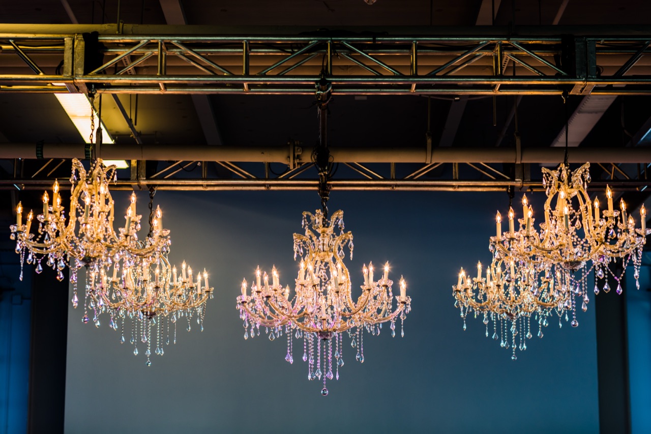 Three ornate crystal chandeliers suspended from overhead truss against a deep-blue draped wall, warmly lit by Impact.