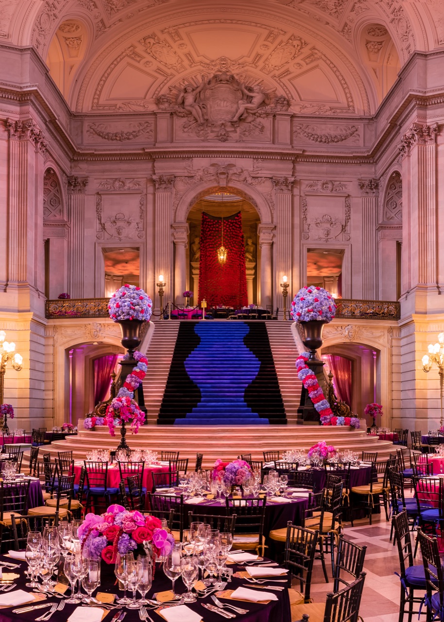 A grand marble staircase dressed with a blue runner and pink florals beneath a red draped backdrop, staged and lit by Impact.
