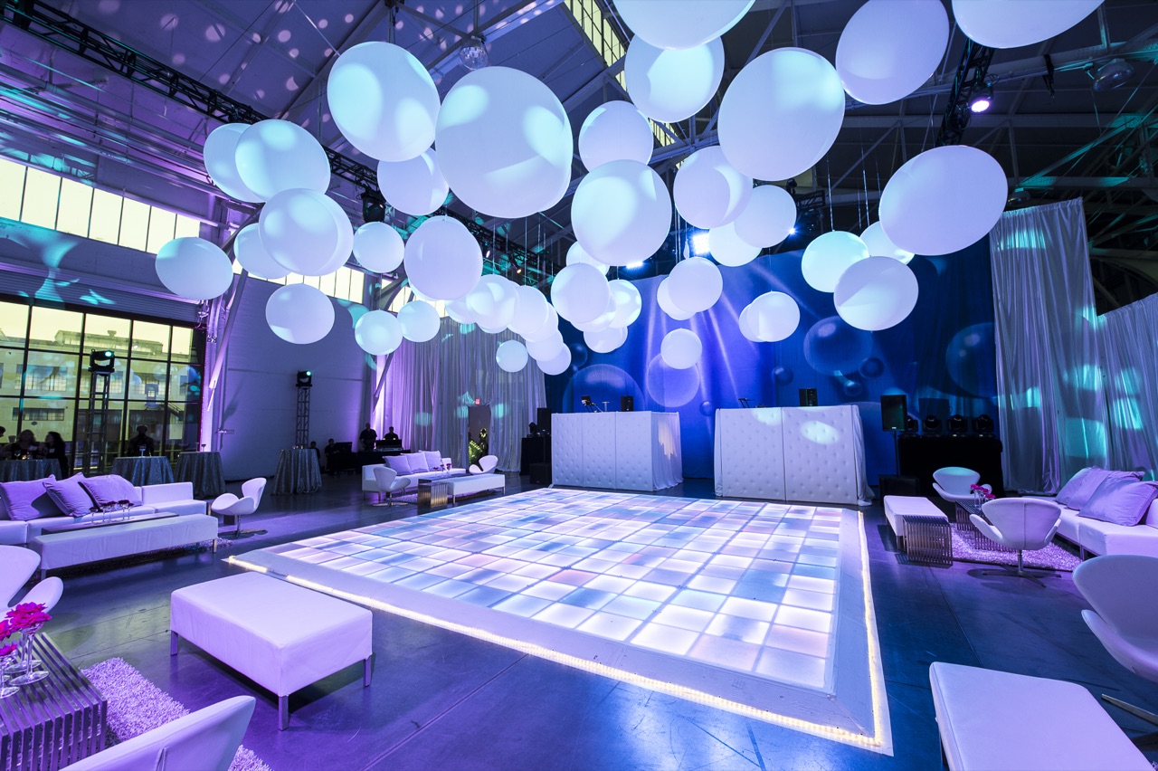 Glowing white orb balloons suspended over an illuminated LED dance floor and white lounge furniture, lit by Impact.