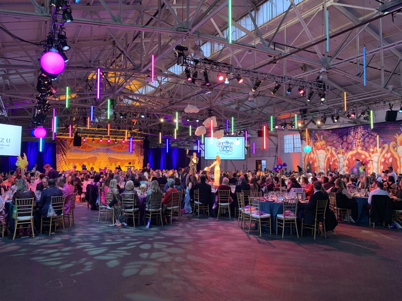 Vertical LED tubes and pink uplighting fill a gala hall of banquet tables facing a lit stage, staged by Impact.